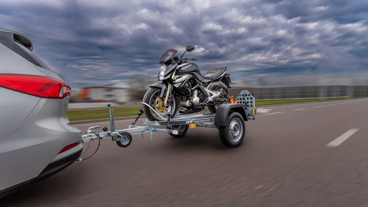 Autoturism argintiu care tractează o remorcă monoax Unitrailer pentru motociclete pe un drum asfaltat, având încărcată o motocicletă neagră Kawasaki bine fixată.