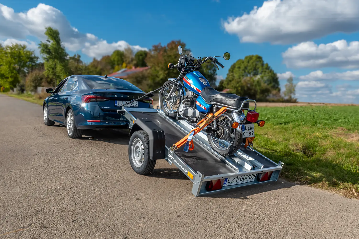 Škoda Octavia albastru închis parcată pe un drum de țară cu o remorcă basculantă Unitrailer pentru motociclete, pe care este fixată cu chingi o motocicletă clasică albastră Honda.
