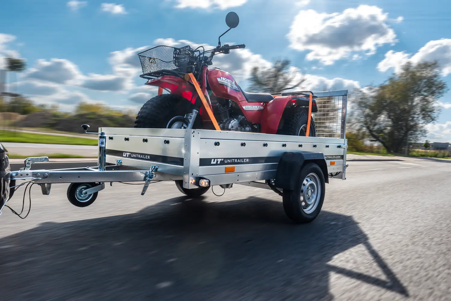 Remorcă monoax cu obloane Unitrailer în mers, care transportă un trike de teren roșu Honda cu coș pe ghidon, ancorat cu chingi portocalii.