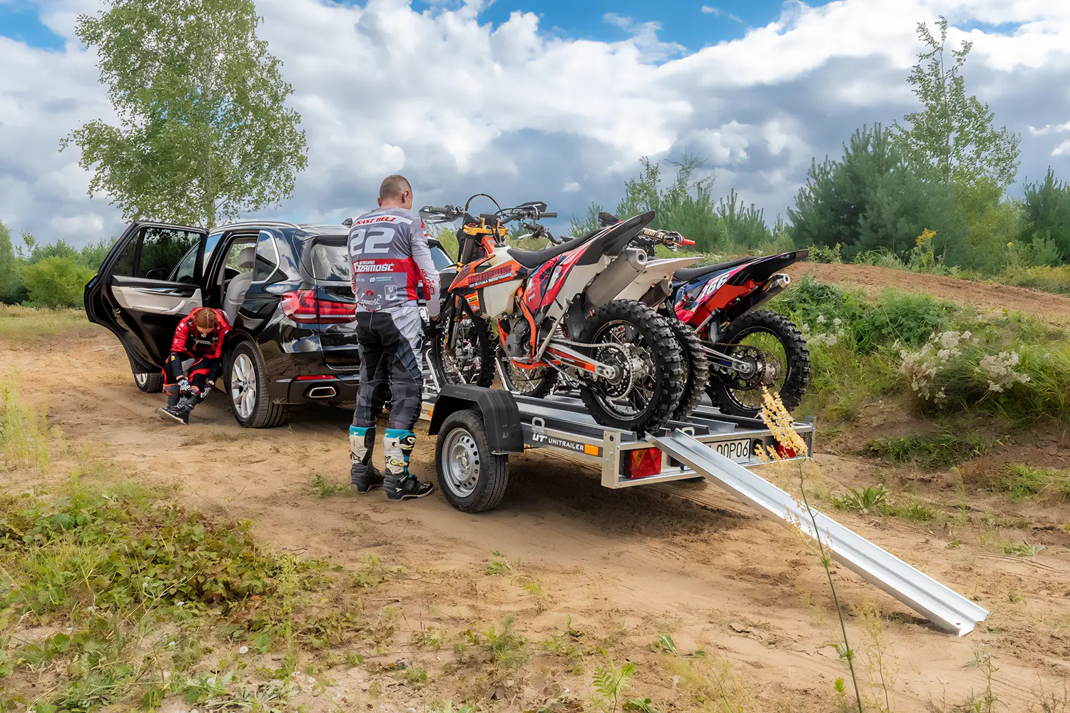 SUV negru parcat pe un traseu off-road cu o remorcă Unitrailer încărcată cu două motociclete de cross, în timp ce doi motocicliști în echipament de protecție se pregătesc lângă rampa de acces.
