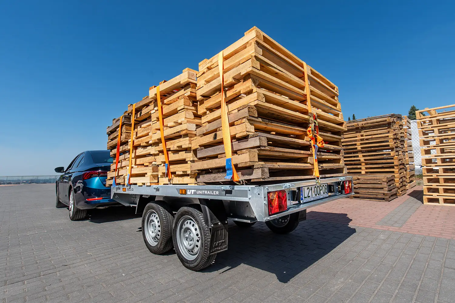 Remorcă de marfă Unitrailer cu două axe, cuplată la o mașină închisă la culoare, încărcată cu o stivă înaltă de paleți de lemn asigurați cu chingi de transport, într-o curte de firmă.