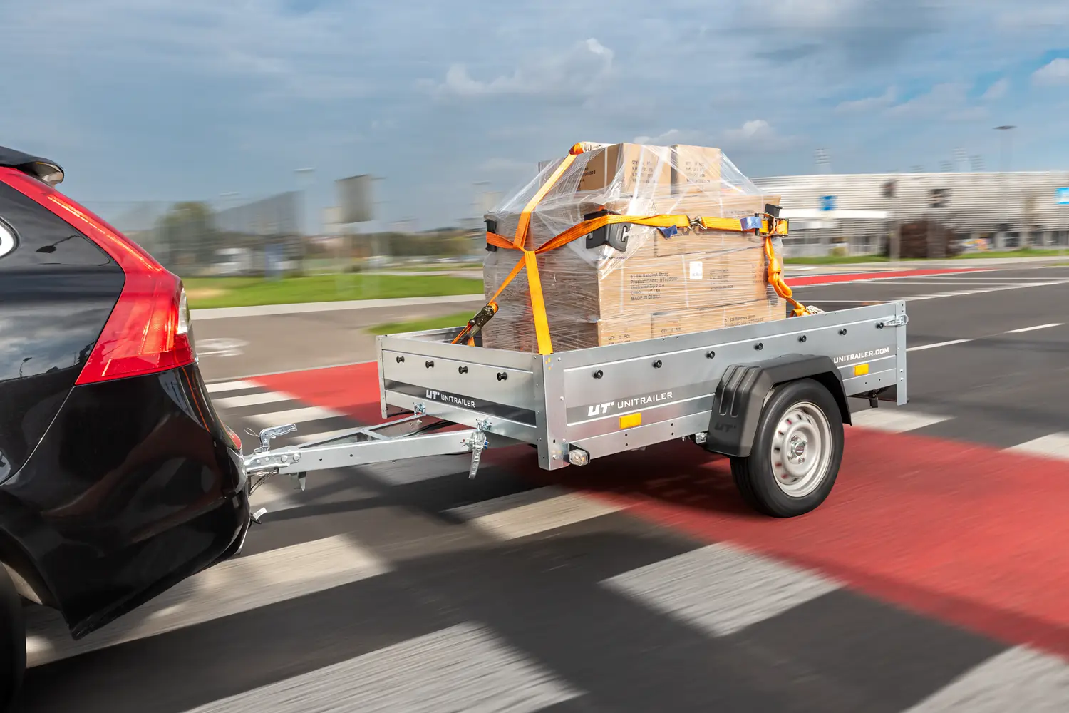 Remorcă de marfă Unitrailer cuplată la o mașină neagră, transportând un palet cu cutii înfășurat în folie stretch și asigurat cu chingi de transport portocalii în timpul deplasării pe drum